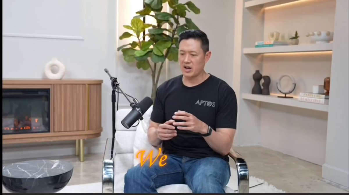 The image shows a man sitting comfortably in a chair while holding a cell phone. The background features a white wall with various decorative items such as vases and potted plants placed around the room. A microphone can be seen, suggesting that this might be an interview or a podcast recording session taking place in the space.