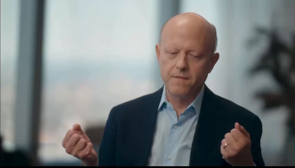 The image features a bald man wearing a suit and tie, sitting at a desk with his hands raised in front of him. He appears to be deep in thought or contemplation as he looks off into space. The background shows a window overlooking an urban cityscape, providing a contrast between the man's serious expression and the bustling city outside.