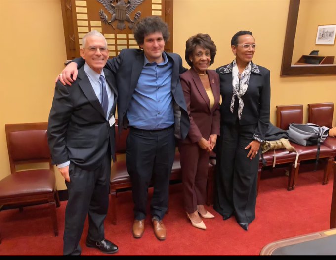 Four people, including Sam Bankman-Fried in a blue shirt and gray suit, pose together for a group photo in what appears to be a formal government setting.