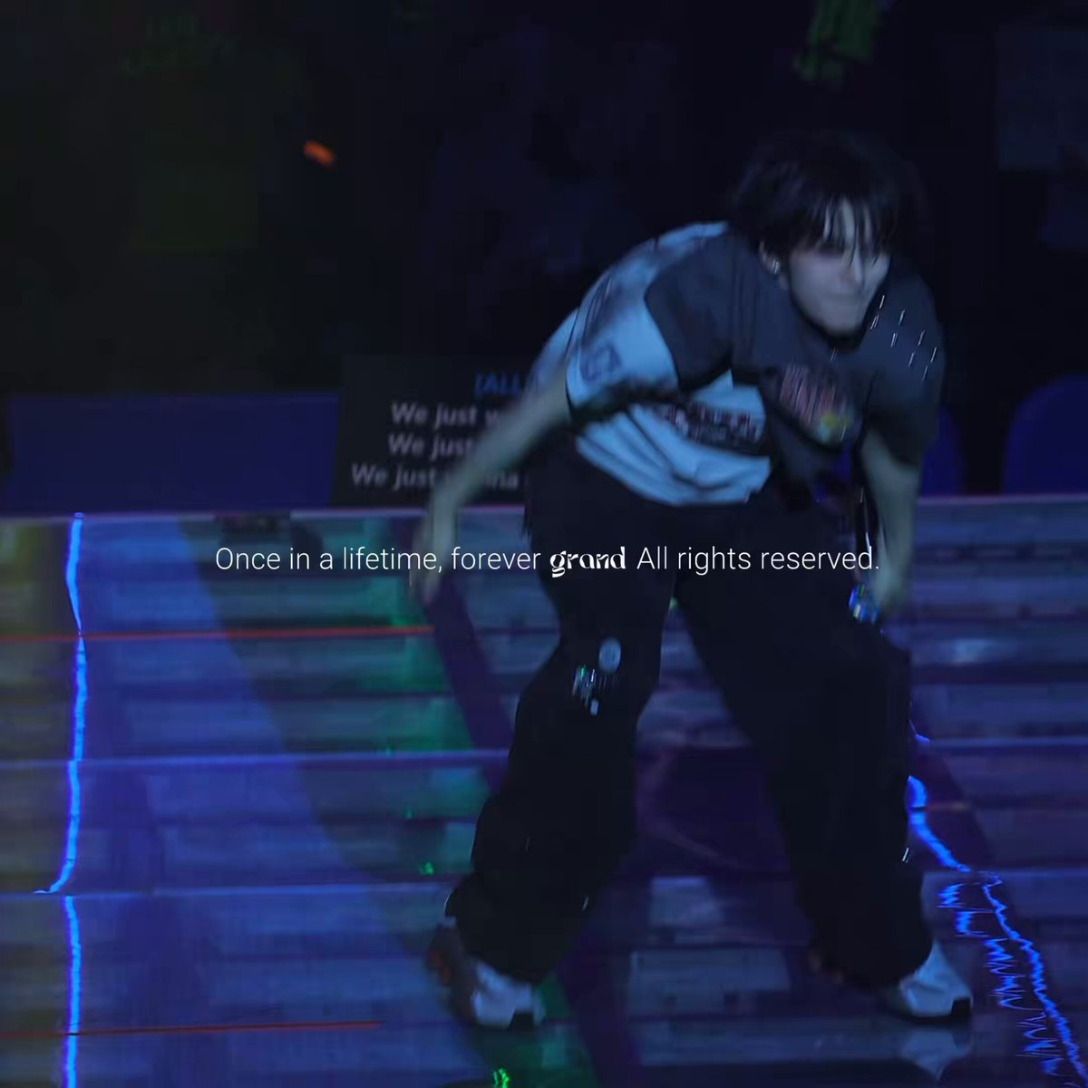 The image features a person wearing black pants, standing on their skateboard with one foot raised while the other remains down. The person appears to be performing a trick or maneuver that requires balance and skill. In the background, there are two chairs visible, suggesting an indoor setting such as a skate park or a similar venue for skateboarding activities.
