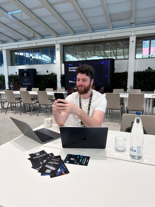 A man with a beard smiles at his phone while seated at a white table with two laptops, water bottles, and business cards in a conference lounge.