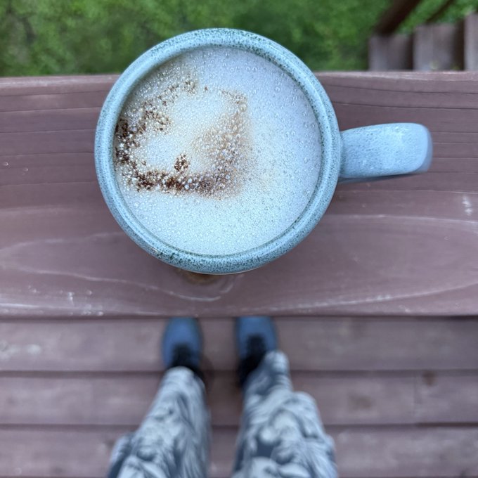 A first-person view of a person's feet in patterned pants and dark shoes standing on a wooden deck, looking down at a speckled blue mug containing a frothy beverage sprinkled with cinnamon or cocoa.