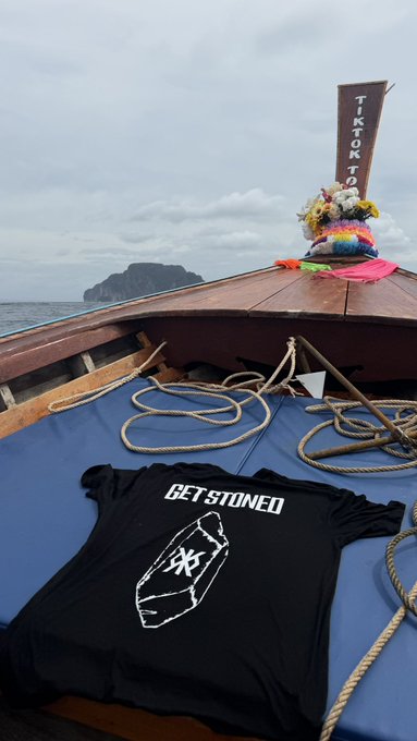 A black t-shirt reading 'GET STONED' with a coffin design laid on the blue deck of a wooden boat, with a 'TIKTOKTO' flag and a floral offering visible on the bow.