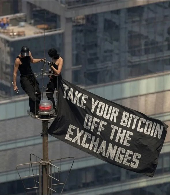 Two people wearing masks stand on a high structure near a building, holding a large black flag with white text reading 'TAKE YOUR BITCOIN OFF THE EXCHANGES'.