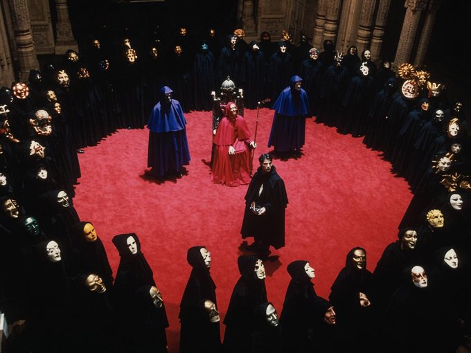 A cinematic scene reminiscent of 'Eyes Wide Shut' featuring masked figures in dark cloaks surrounding a red-carpeted ritual area where a man in black stands before robed figures holding ceremonial items.