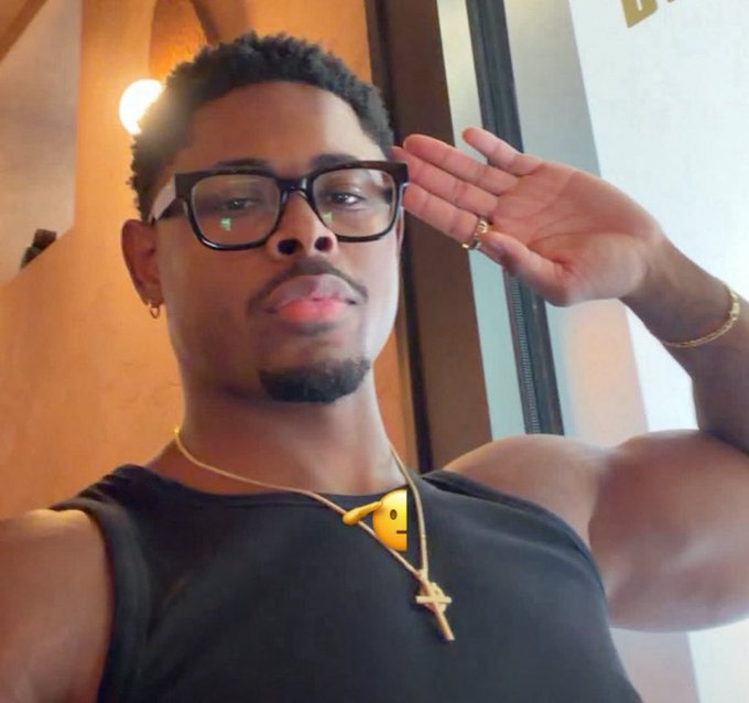 A selfie of a young man wearing glasses, a black tank top, and gold necklaces, saluting with his hand near his forehead.