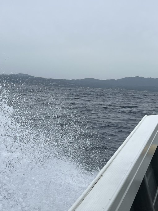 The image shows a boat cutting through choppy water, creating a large spray of white foam behind it. The boat's wake is visible as it moves across the ocean. In the background, there are mountains that add depth and beauty to the scene. The photo appears to be taken from inside or on the boat itself, capturing the motion and energy of the moment.