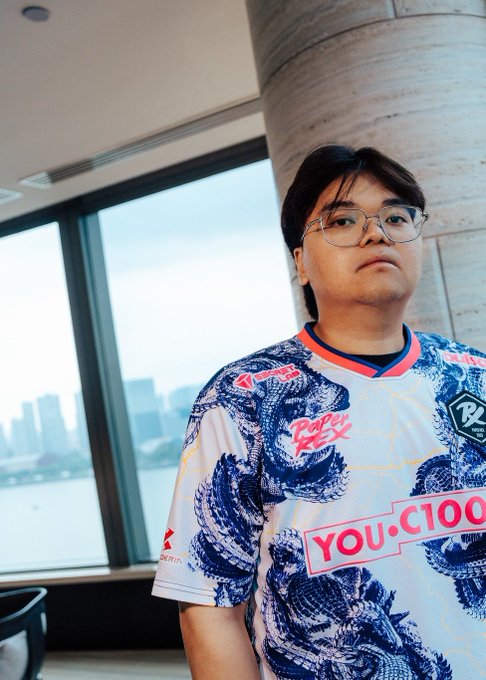 A young man with dark hair and glasses poses indoors near a window, wearing a white esports jersey with blue dragon designs featuring logos for "Paper Rex," "Singapore Airlines," and "YOU.C1000." The jersey reflects his role as a member of the Paper Rex VALORANT team, matching the post's quote where he critiques his own performance in a loss to LOUD.