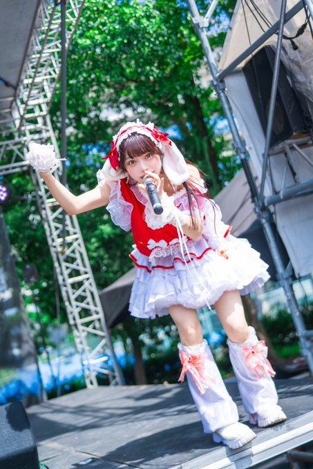 A young female performer on stage singing into a microphone, wearing a red and white frilly idol costume with a bonnet and tall boots.