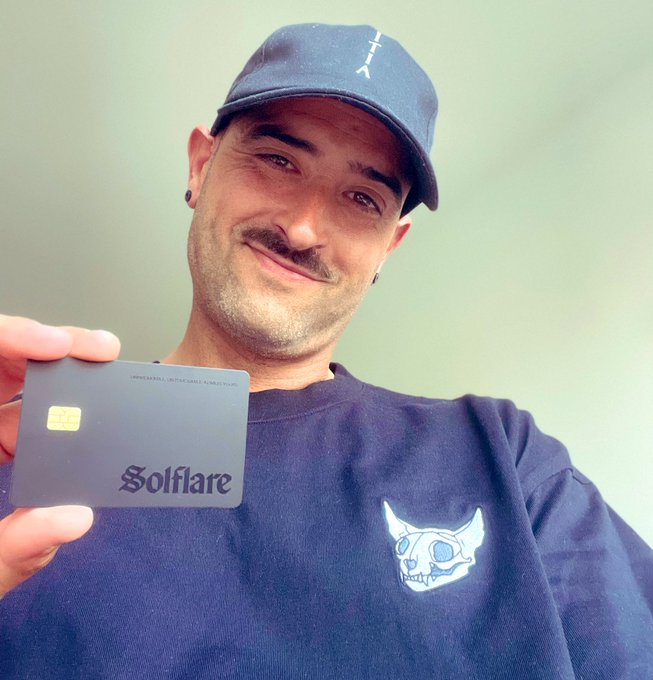 A man wearing a navy cap and a black t-shirt featuring a horned skull logo smiles while proudly holding up a dark gray Solflare crypto wallet card, perfectly capturing the post's sentiment of "Unbreakable, Untouchable. Always" with the branded card displaying the "Solflare" name and a visible EMV chip.