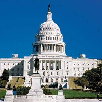 The United States Capitol Building with its iconic white dome stands against a clear blue sky, featuring a statue in the foreground.