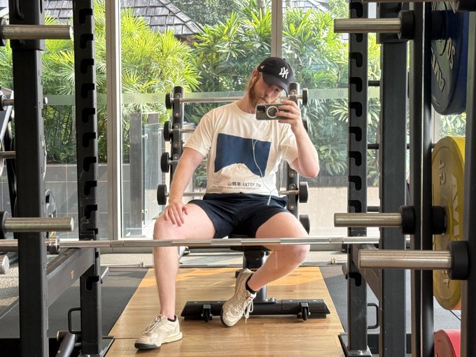 A man in a New York Yankees cap, white t-shirt, and black shorts sits on a bench in a gym, taking a mirror selfie with his phone.