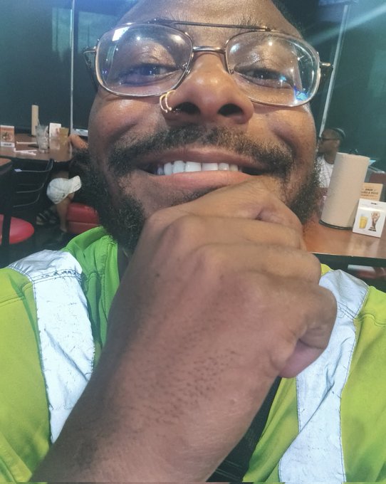 A man in a green shirt and wire-framed glasses smiles broadly while resting his chin on his fist, posing for a selfie in what appears to be a restaurant setting. The joyful expression visually reinforces his celebratory "#Day559" ApeCoin ($APE) milestone post, showing his enthusiasm as he counts down to the token's all-time high.
