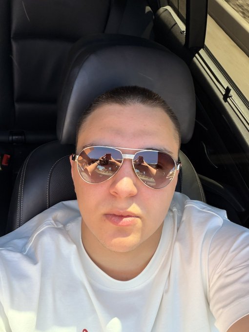 The image features a man wearing sunglasses, sitting inside a car with his head resting against the back seat of the vehicle. The man appears to be posing for a photo or selfie, as indicated by the caption "Gm! Say It Back and Get $$$$ Drop Your Wallets!".