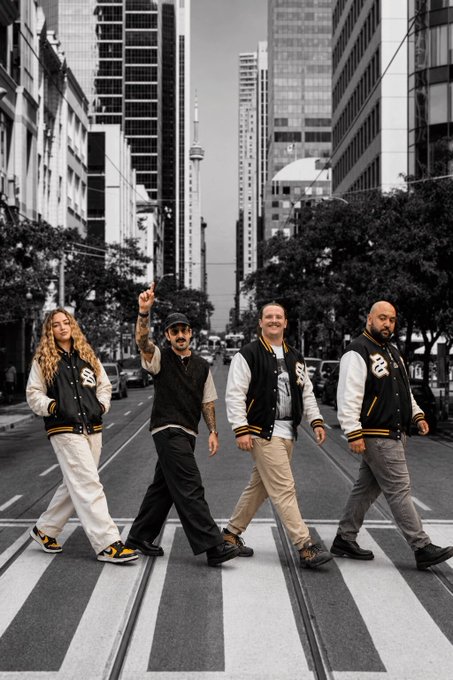 Four people wearing matching black and white varsity jackets cross a city street in a posed photo, recreating a Beatles Abbey Road-style crossing with urban skyscrapers and a tower in the background. The image serves as a stylized farewell tribute to departing team members @mauricedotxyz, @leclop_sol, @devmajesty, and @satoshitriangle, symbolizing the group walking off together in different directions as the organization evolves.