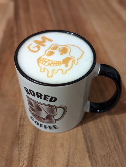 A white and black mug labeled 'BORED COFFEE' sits on a wooden surface, containing a latte with 'GM' and a skull design created in the foam.