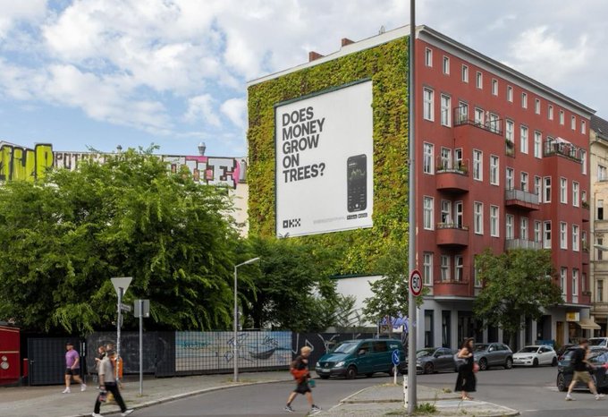 A street view of a red building covered in green ivy with a large billboard asking 'DOES MONEY GROW ON TREES?' and featuring an OKX cryptocurrency logo.