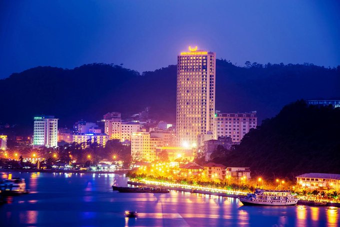 The image shows a city skyline at night, with tall buildings lit up against the dark sky. The tallest building has its lights turned on, illuminating the surrounding area. A boat can be seen floating near the shoreline of a river or lake, adding to the overall ambiance of the scene. The combination of the illuminated buildings and the natural water body creates an enchanting atmosphere in the cityscape.