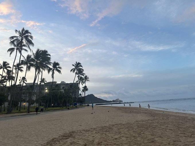 The image shows a beautiful beach scene at sunset, with palm trees lining the shoreline. There are several people enjoying their time near the water's edge or walking along the sandy beach. The sky above them is filled with clouds, creating a picturesque backdrop for this serene setting. In the background, there is a hotel building visible, indicating that this location might be a popular spot for tourists and locals alike to relax and unwind during their vacations.