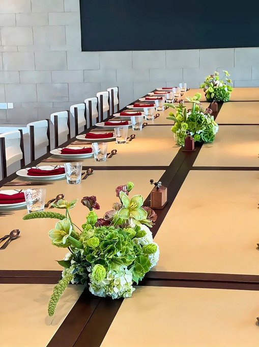 An elegantly set long banquet table featuring cream-colored runners, dark wood trim, red napkins, water glasses, and lush green and white floral centerpieces with a dark screen on the wall in the background.