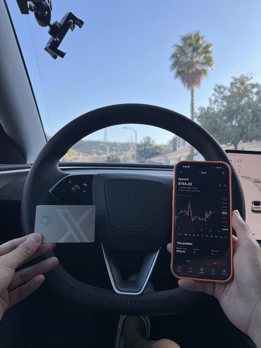 A person holds a Tesla-branded credit card in one hand and a smartphone displaying the SpaceX stock chart ($154.42) in the other while sitting behind the steering wheel of a Tesla vehicle.