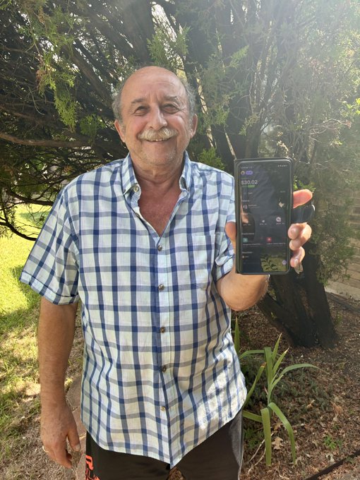 A smiling older man with a mustache wearing a blue and white plaid short-sleeved shirt, standing outdoors in front of a tree and proudly holding up a smartphone displaying a trading app showing '$33.66'.
