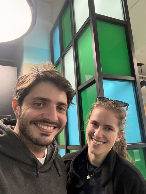 A smiling man and woman take a selfie together in front of a colorful stained-glass style window with blue and green panels.