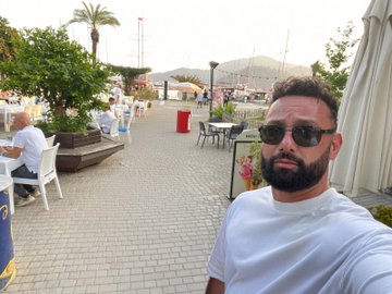 A man in sunglasses and a white t-shirt takes a selfie outside a coastal promenade with restaurants and palm trees. The image accompanies "Day 2 of posting a selfie until $HYPE is 3 digits," showing him outdoors on what appears to be a vacation, still waiting for the token to reach triple digits.