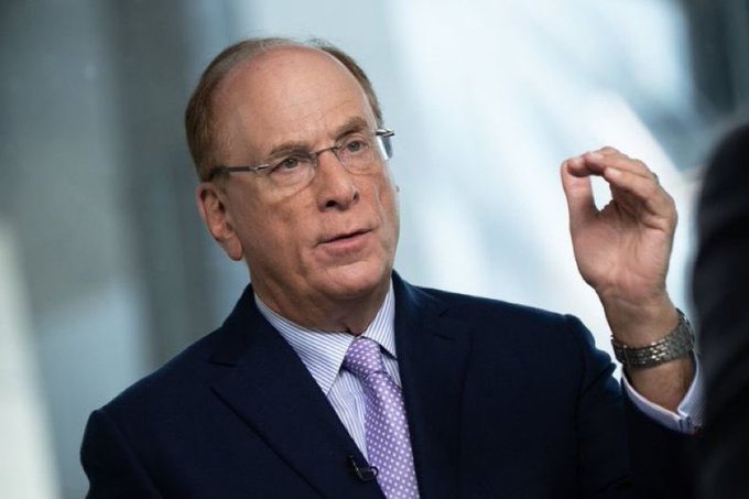 Larry Fink, CEO of BlackRock, is shown speaking and gesturing with his hand, wearing a dark suit and purple tie, likely in an interview setting discussing the volatile performance of the company's $IBIT Bitcoin ETF, which has seen early investors shift from significant gains to substantial losses amid the crypto market's recent downturn.