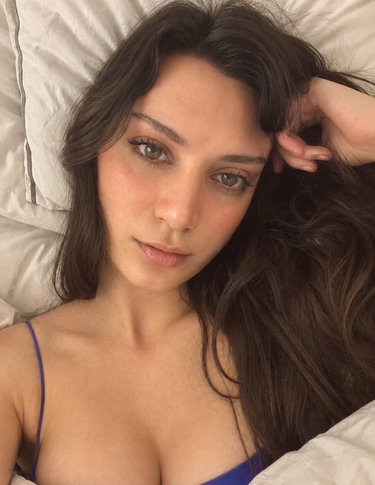 A close-up selfie shows a young woman with long, wavy dark brown hair and brown eyes lying on white bedding, wearing a blue top with thin straps.