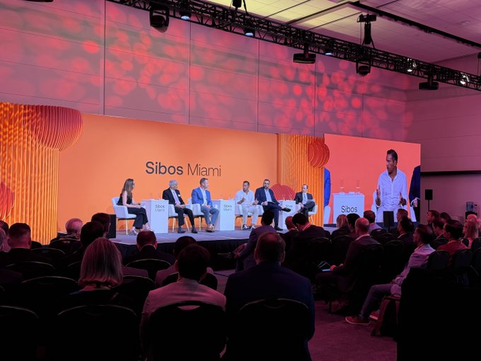 A panel discussion at the Sibos Miami conference, with speakers seated on a stage under pink lighting in front of an audience.