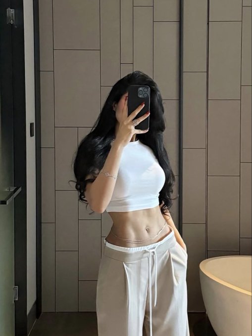 A woman with long dark hair takes a mirror selfie in a bathroom, wearing a white crop top and beige sweatpants, holding a phone that covers her face.
