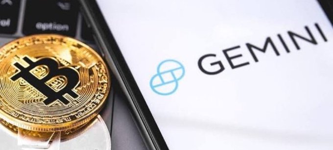 A physical Bitcoin token rests beside a smartphone displaying the Gemini cryptocurrency exchange logo, visually illustrating the post's report that Gemini has expanded its Bitcoin treasury holdings to 5,528 BTC, including a recent purchase of 1,689 BTC.