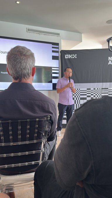 A speaker in a purple shirt addresses an audience at the OKX creator retreat, with the OKX logo visible on the screen and backdrop behind him, while the back of an attendee in a dark shirt is seen in the foreground. The image captures the event setting referenced in the post, showing the intimate gathering where product feedback was discussed with the OKX team.