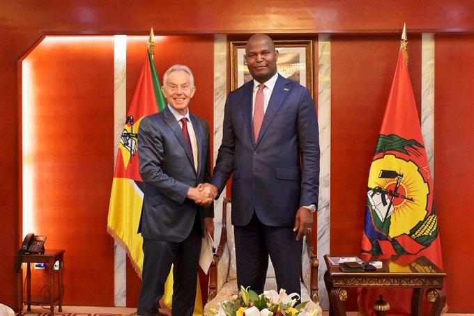 Two men in suits shake hands in a formal setting between the flags of Mozambique and what appears to be a regional emblem, with a framed portrait visible in the background.