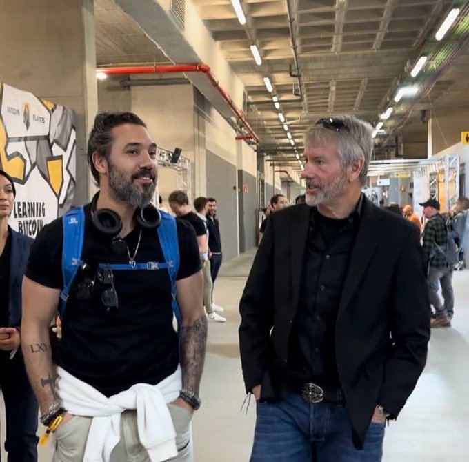 The image shows two men standing next to each other in a hallway or an indoor space with a large open area. One of them has a backpack, while the other man is wearing glasses. They appear to be engaged in conversation, possibly discussing something related to their surroundings. The background features a wall adorned with various posters and signs, which adds to the overall atmosphere of the scene.