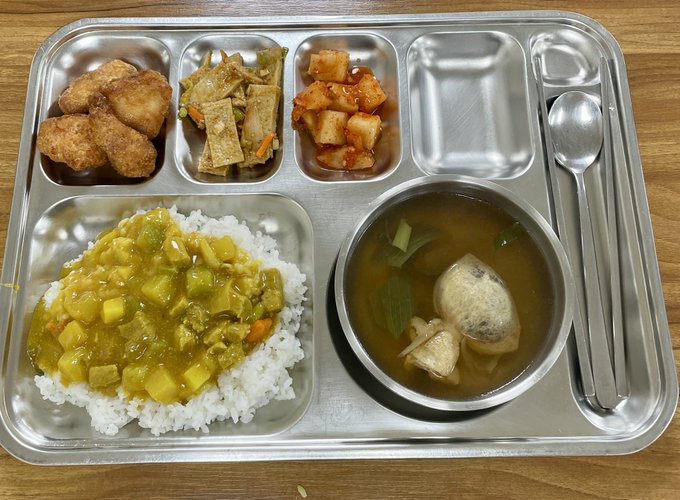 A Korean-style metal cafeteria tray holding a school lunch with curry rice, soup with dumplings, fried chicken, braised fish cakes, and kimchi, accompanied by a spoon and chopsticks.