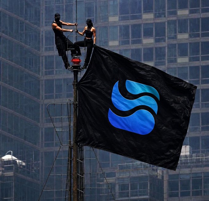 Two people in black clothing and face coverings stand atop a pole, unfurling a large black flag featuring the blue Sui blockchain logo against a city skyscraper backdrop.