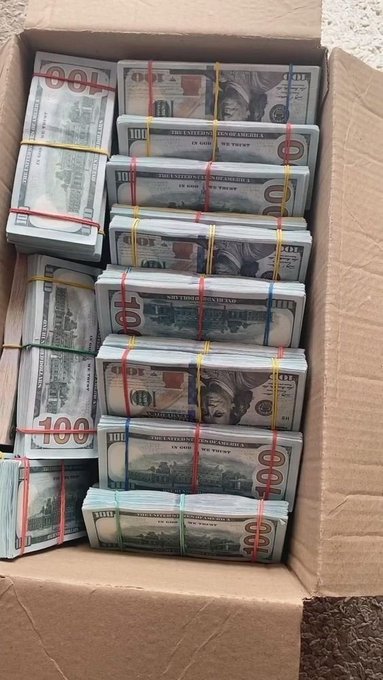 A cardboard box is filled with stacks of $100 US bills secured with colorful rubber bands, representing the $5,000 cash prize offered in the giveaway where participants pick a number between 20–72 to win one of 20 randomly selected spots.