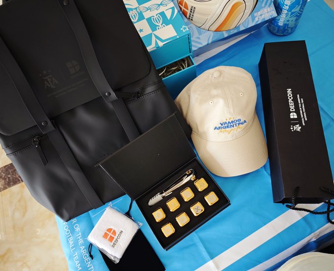 Argentina football team merchandise is displayed on a blue cloth, including a black backpack, a cream cap reading 'VAMOS ARGENTINA', a black 'DEPCOIN' box, a pin collection, and a 'WE ARE ARGENTINA FOOTBALL TEAM' badge.