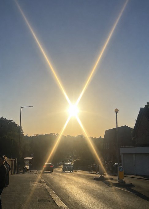 A dramatic X-shaped sunburst dominates the London skyline, with the setting sun casting bright rays that resemble an "X" over a quiet street with a few cars and pedestrians. The striking cross pattern in the sky playfully echoes the "XRP" ticker symbol referenced in the caption, suggesting a celestial "signal" for the cryptocurrency.