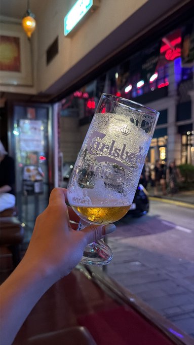 The image features a person holding a glass of beer at an outdoor restaurant or bar. The beer appears to be a pilsner style, which has a golden color that stands out against the dark background. There are other people present in the scene as well, suggesting that it is a lively and social atmosphere.
