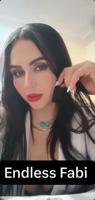A woman with long dark hair and bold makeup (red lips, smoky eyes) wearing a light-colored robe and layered necklaces, with the caption "Endless Fabi" displayed at the bottom.