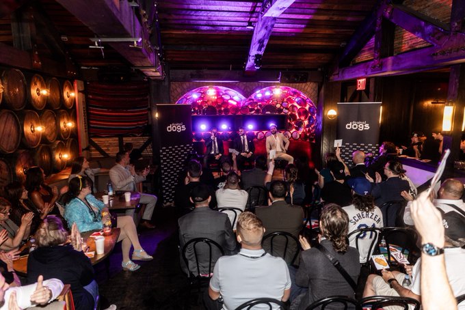 An audience sits in a dimly lit venue with wooden barrels, watching a panel discussion on stage with 'dōgs' branding displayed on the backdrop.