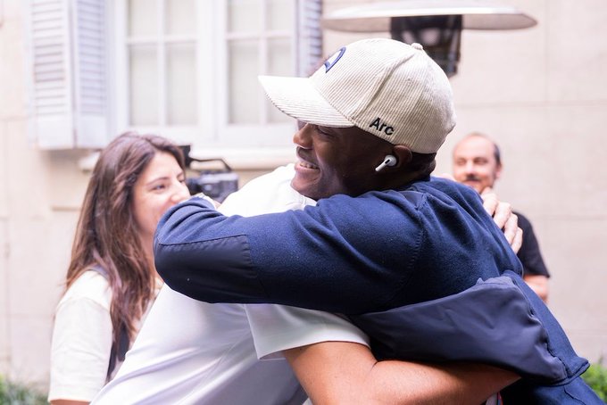 iced coffee and a white hat with blue lettering are present, accompanied by two individuals sharing a warm embrace against a beige wall backdrop.