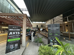 A photograph of a building entrance with a 'WELCOME AXTS' banner and an event sign featuring Batman artwork, with attendees gathered near the doors.