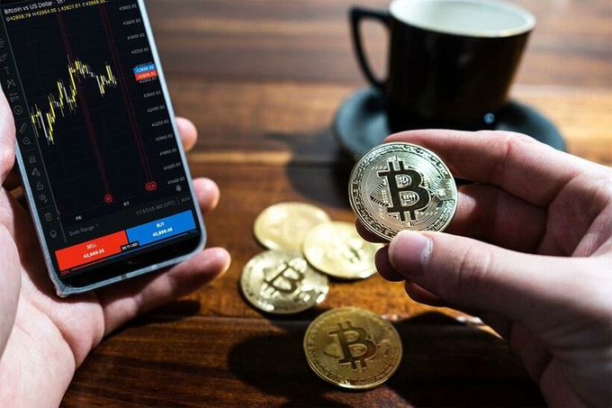 A hand holding a gold Bitcoin token in front of a smartphone displaying a BTC/USD trading chart from a US exchange, with additional Bitcoin tokens scattered on a wooden table and a coffee cup in the background.