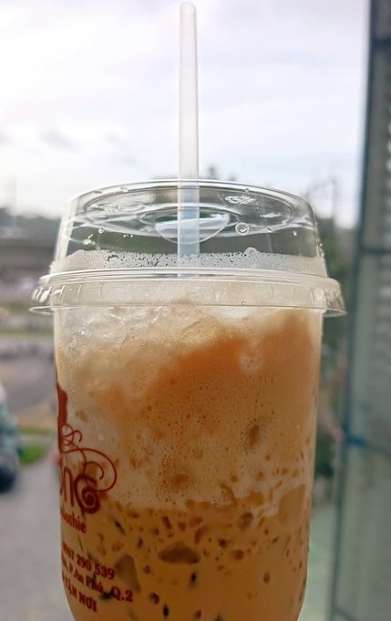 A close-up photo of an iced Thai milk tea in a clear plastic cup with a dome lid and straw, showing partial text 'Cha...', '...290 539', and '...A.Q.2'.