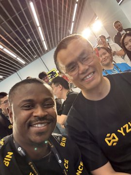 Two smiling men pose for a selfie, one wearing a black "BINANCE" lanyard, alongside other team members in an indoor venue, with the post referencing the Binance legends behind the world's first super app and playfully suggesting they would burn 70% of a meme token's supply rather than sending it to CZ or Giggle Academy.