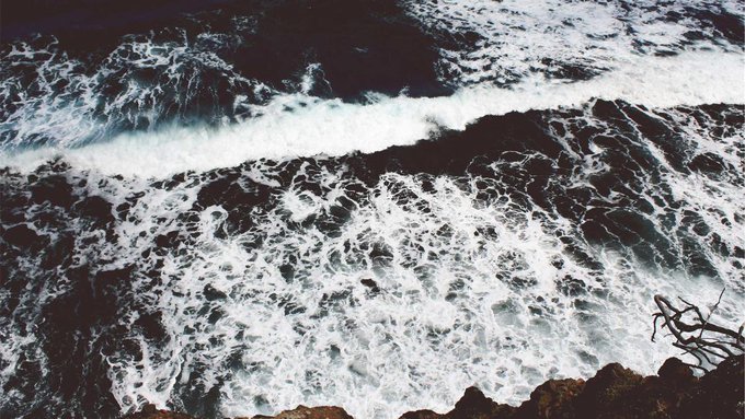 An aerial view photograph of dark ocean waves crashing against rocky shores, creating dramatic white foam patterns across the water.